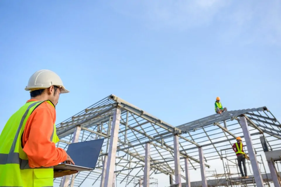 Asian Male Construction Engineer Holding A Laptop Standing At A House Construction Site Controlling And Ordering Workers Building Houses And Steel Roof Trusses.